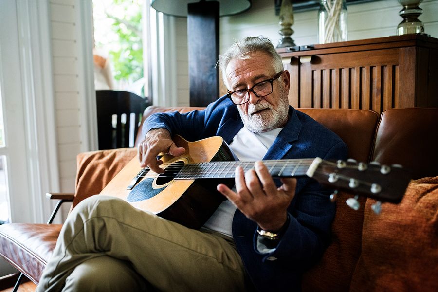 A photo of an older man, sat on a couch, playing an acoustic guitar.